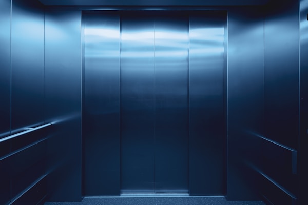 Closed elevator doors in a modern, metallic elevator interior with blue lighting and handrails on both sides.