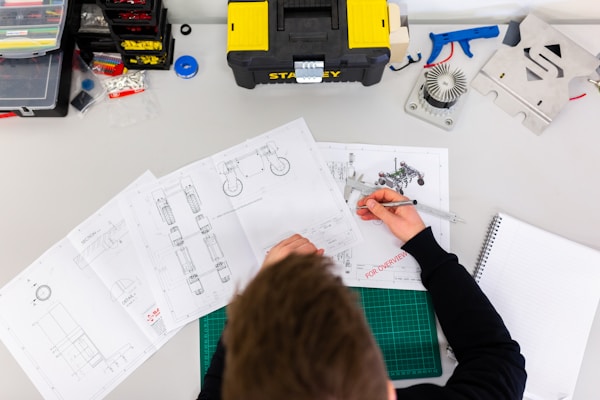 Person reviewing technical drawings at a desk with tools, a caliper, and engineering equipment visible, viewed from above.