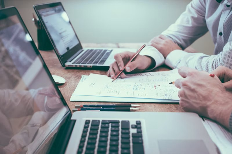 Two people sit at a desk with laptops, reviewing documents and taking notes with pens.