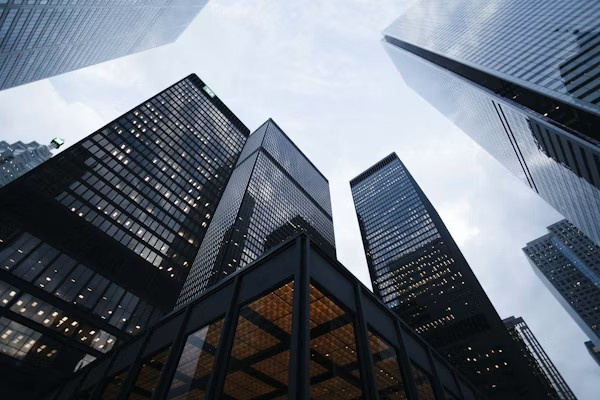 Tall modern skyscrapers seen from below against a cloudy sky, featuring glass and steel facades reflecting light.