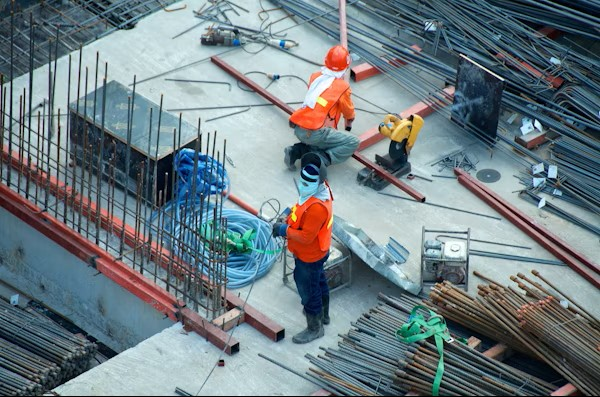 Two construction workers in safety gear work amid steel rods and equipment on a concrete structure at a building site.