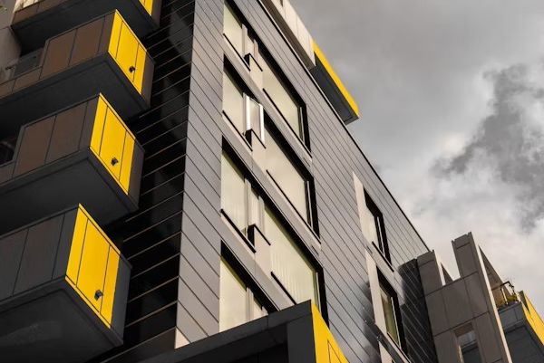 Modern apartment building with gray facade and yellow balconies under a cloudy sky, viewed from a low angle.