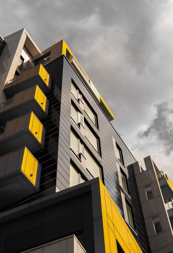 A modern multi-story building with gray and yellow exterior panels, balconies, and large windows, set against a cloudy sky.